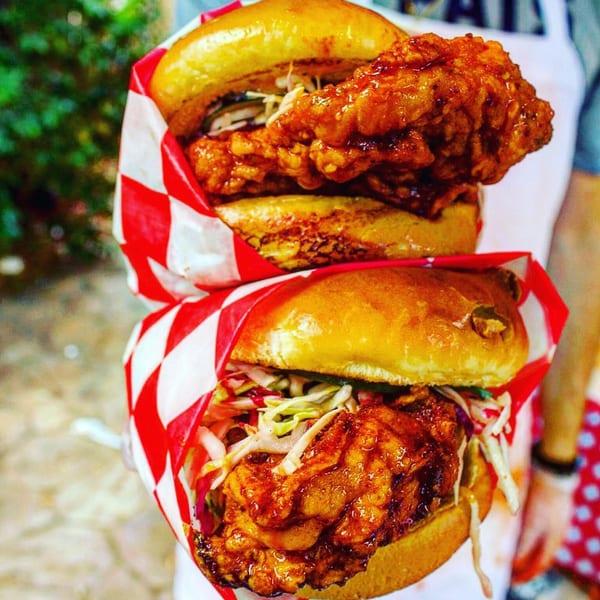 Two 2 Neighbor's fried chicken sandwiches, with slaw and sauce, wrapped in red and white checkered paper.