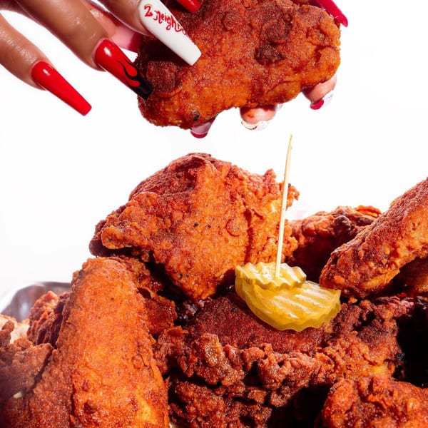 A hand with long red and white nails holding a piece of 2 Neighbor's fried chicken over a pile of chicken.